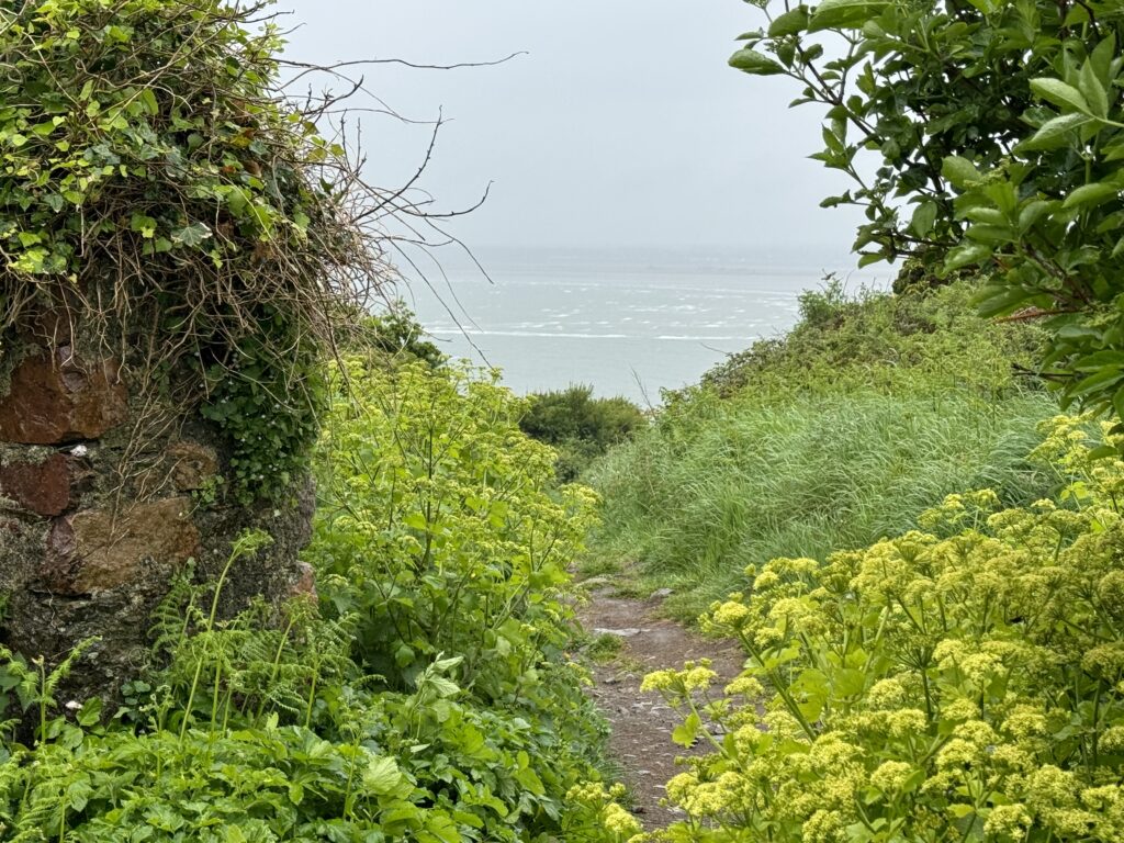 A path passing by a stone pillar on the left. there is a grassy bank on the right with a grey sea and grey clouds in the background.