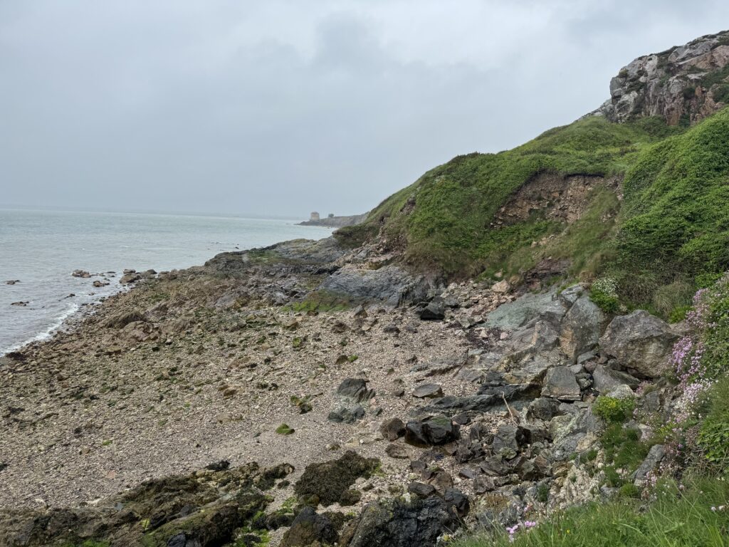 A rocky beach with a grassy embankment leading up to a cliff on the right. A Martello tower can be seen in the distance. The sea is on the left with grey skies overhead.
