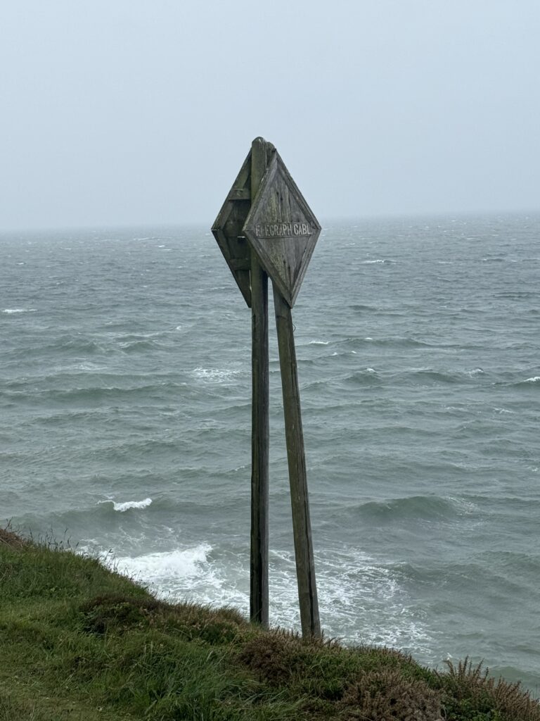 A closer view of the signpost shows that it reads “Telegraph Cable”. The photograph is from right at the ends of the cliff with Andy a small amount of ground viewable. A choppy sea makes up the background with grey skies overhead.