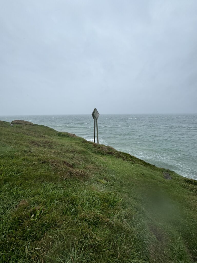 A tall signpost at the edge of a cliff. A grassy bank on the left with a choppy sea on the right. Grey skies overhead.