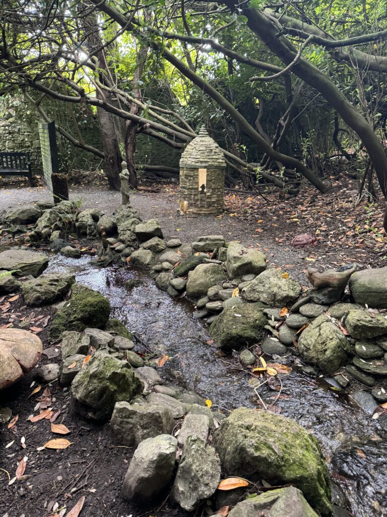 A stream with banks of stone on either side is in front of small round stone structure within the fairy garden in Belvedere. Trees grow behind the stone structure.