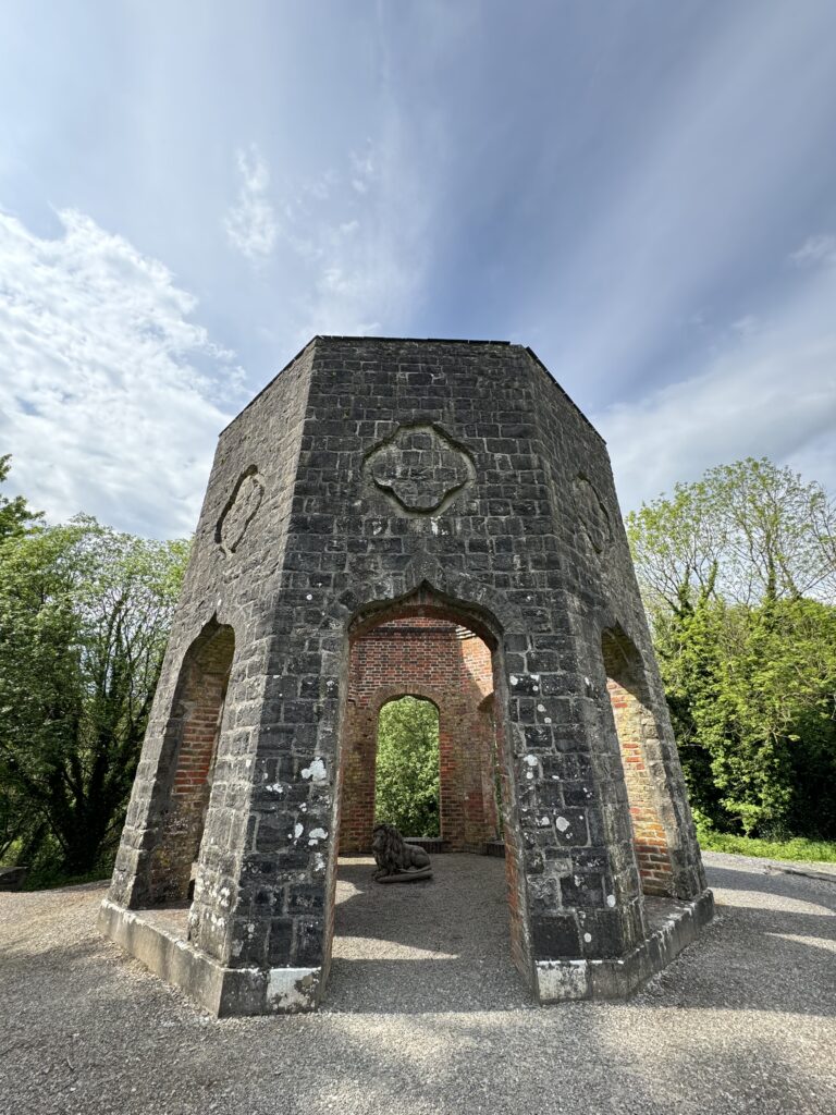 An Octagonal Gazebo built on a raised structure. A statue of a lion can be seen within the Gazebo. Blue sky overhead.