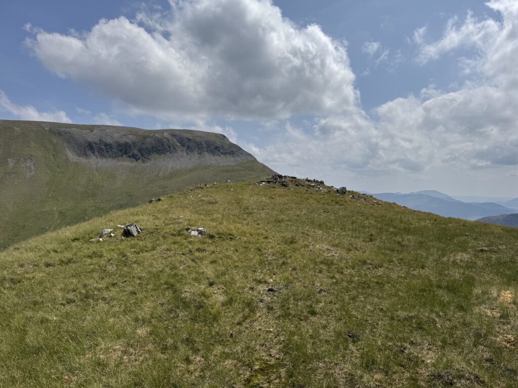 Grassy summit ridge with scattered rocks while climbing Barrclashcame, with dramatic cliffs and cloud-filled skies in the distance.