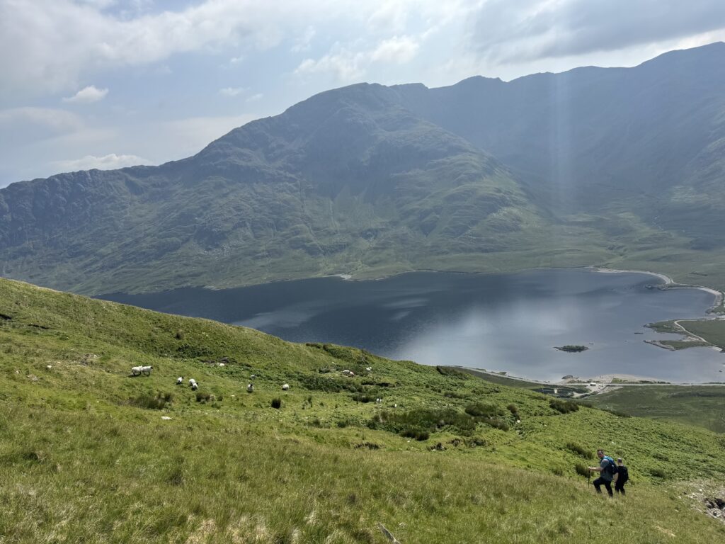 Hiker descending steep grassy slope  with Doolough Lake and Mweelrea Mountains in the background.