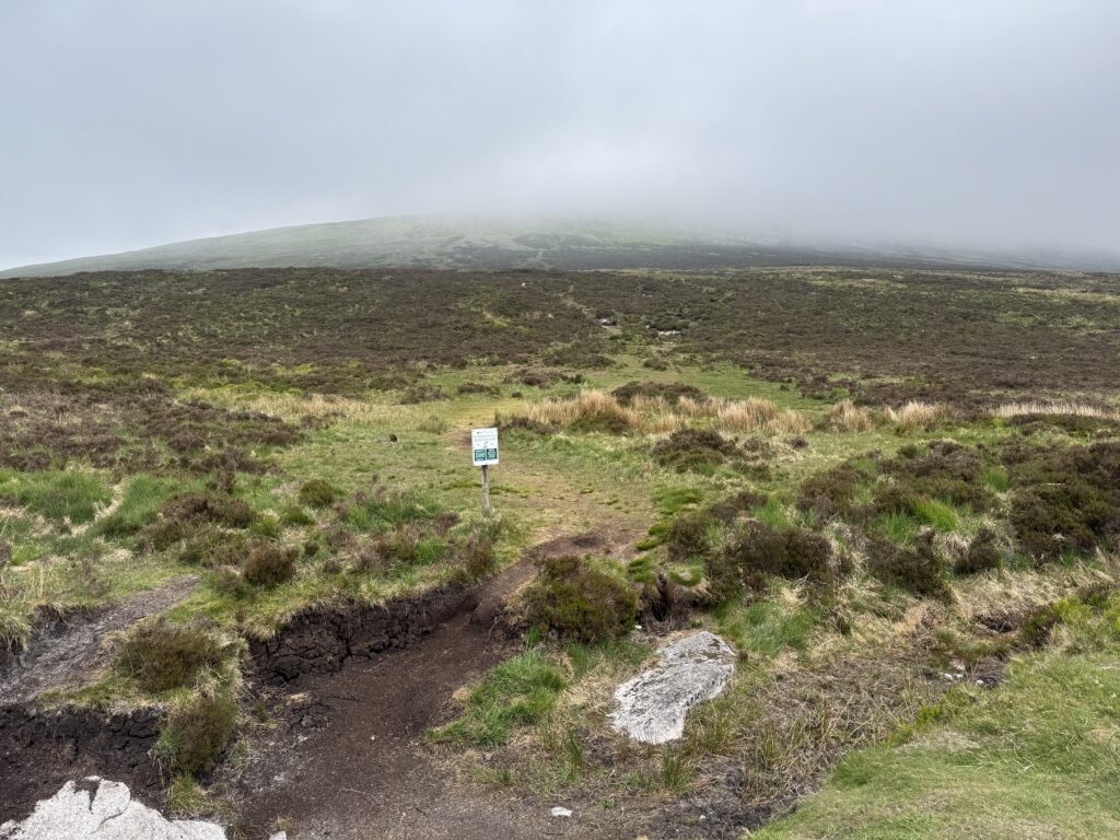 A rough path can be faintly seen snaking up the slope of a mountain. The top of the mountain is covered by cloud.