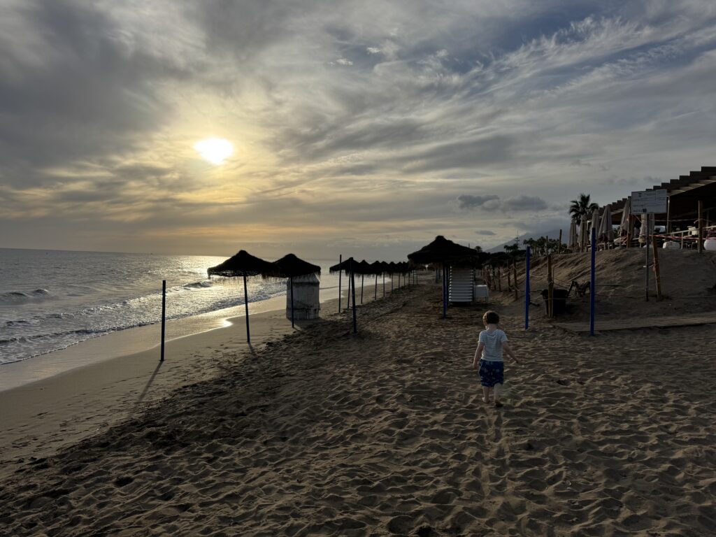 The Beach! We spent a lot of time here during our week in Elviria with a 3 year old.
A small boy walks towards sun loungers on a shady beach with the sun setting in the background.