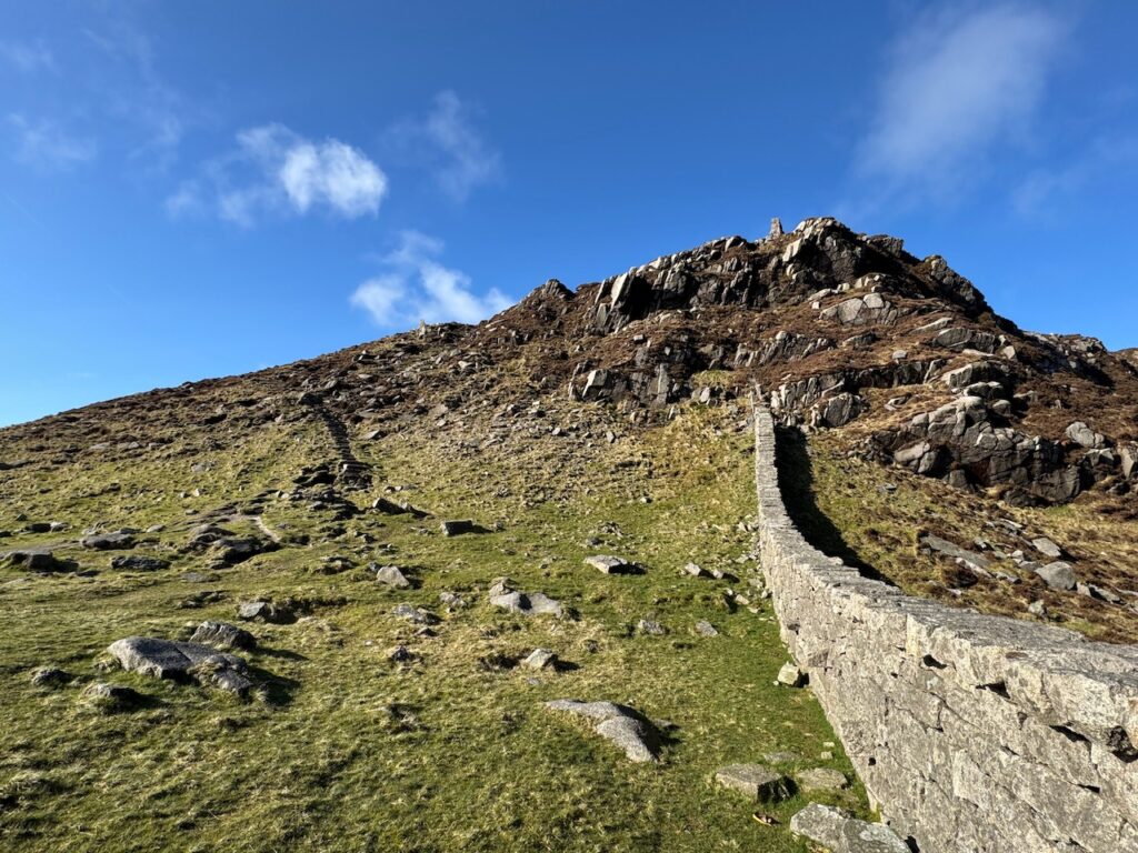 Mourne Wall at Hare’s Gap along the route of the Slieve Bearnagh hike. The wall fades into the steepness of the cliff. A path cuts into the cliff on the left. Blue skies overhead