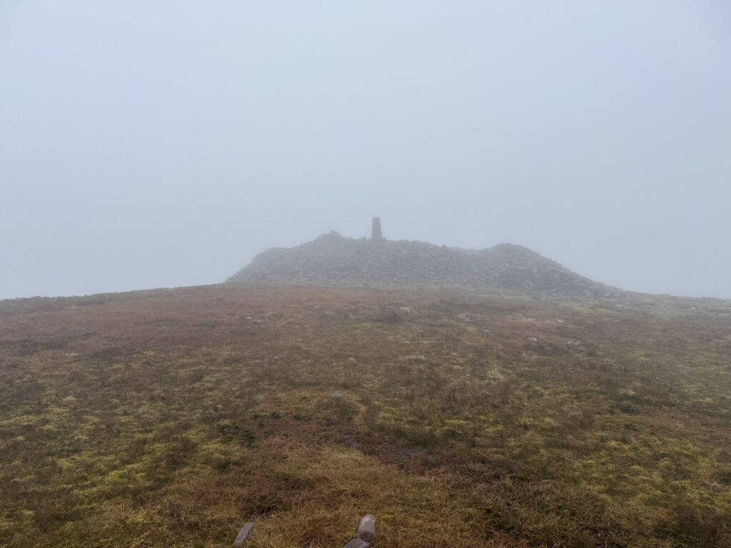 Temple Hill Walk approaching the stone summit cairn and trig pillar through dense mist and low cloud.