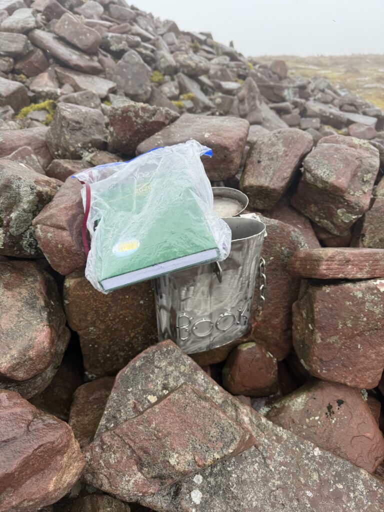 Hikers’ guest book in a waterproof bag beside its metal container among the wet stones of the summit cairn.