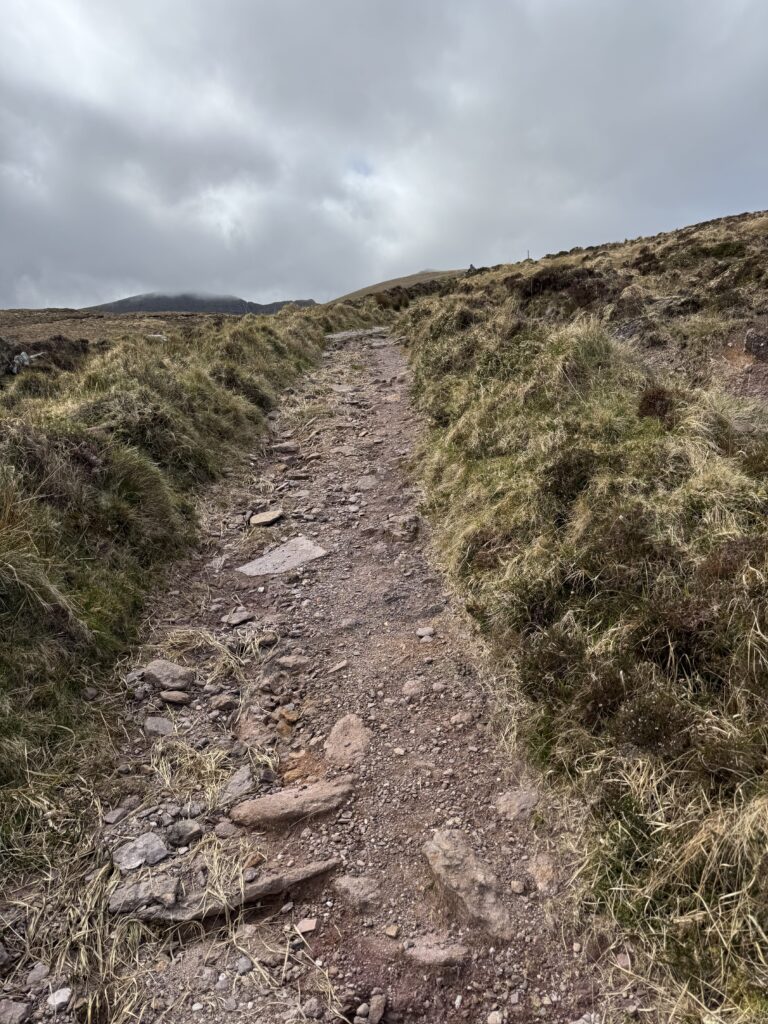 Steep rocky mountain path climbing through grassy slopes towards the cloud-covered summit of Masatiompan.