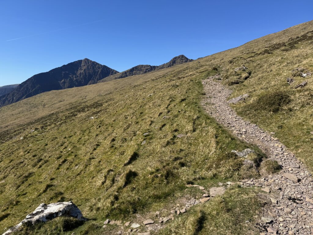 Mountain path climbing across open grassland towards the steep rocky cliffs of the Brandon Mountains.