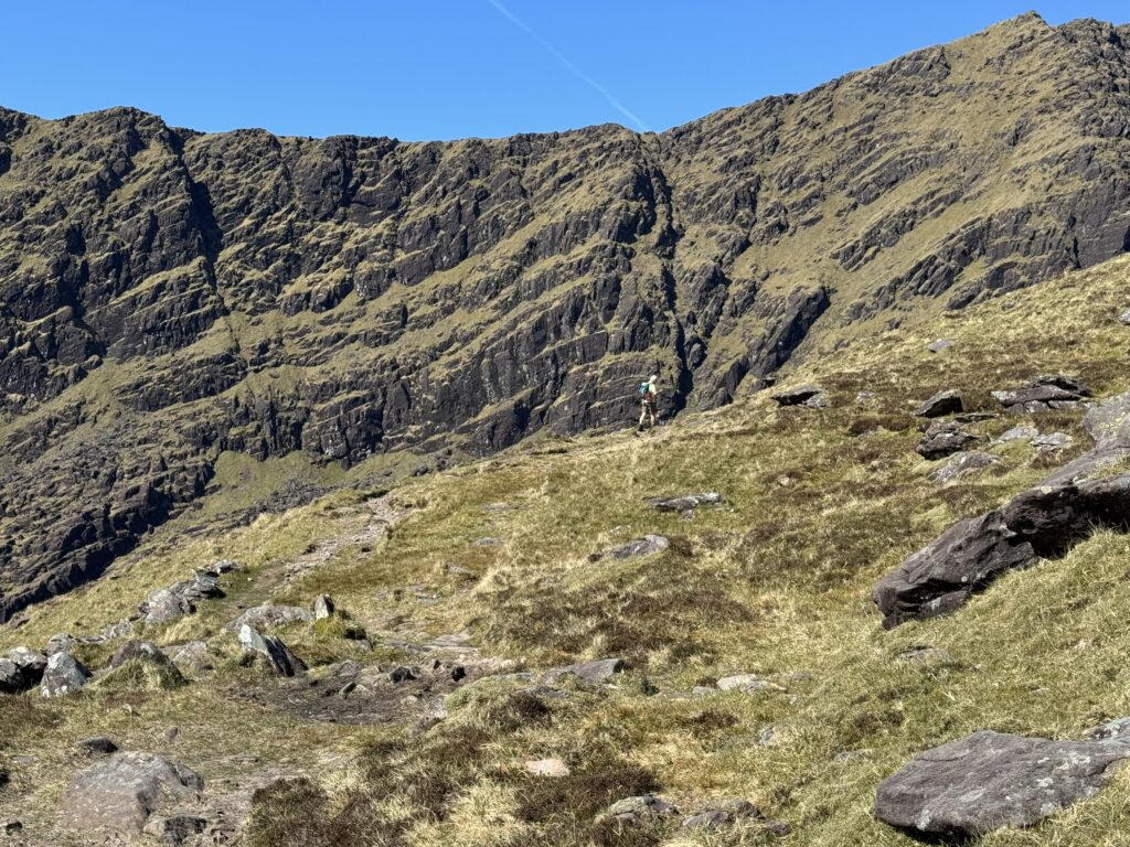 Hiker following the Mount Brandon Loop beneath the towering rocky cliffs of the Faha corrie on the Dingle Peninsula.