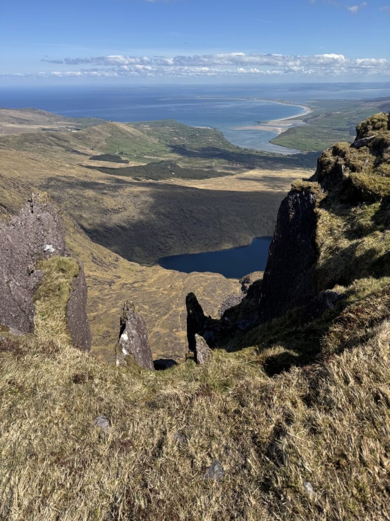 View between steep rocky cliffs towards a mountain lake, with Brandon Bay and the Atlantic Ocean beyond.