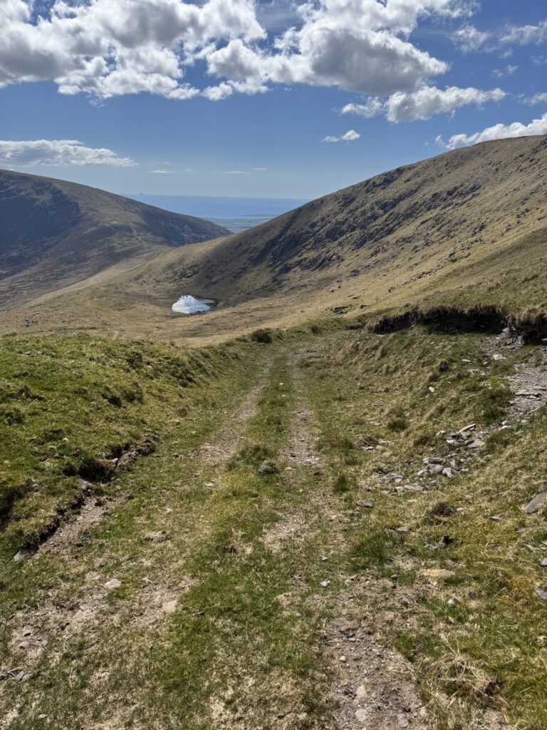 Grassy mountain track descending towards a small lake between the surrounding ridges, with the Atlantic visible beyond.