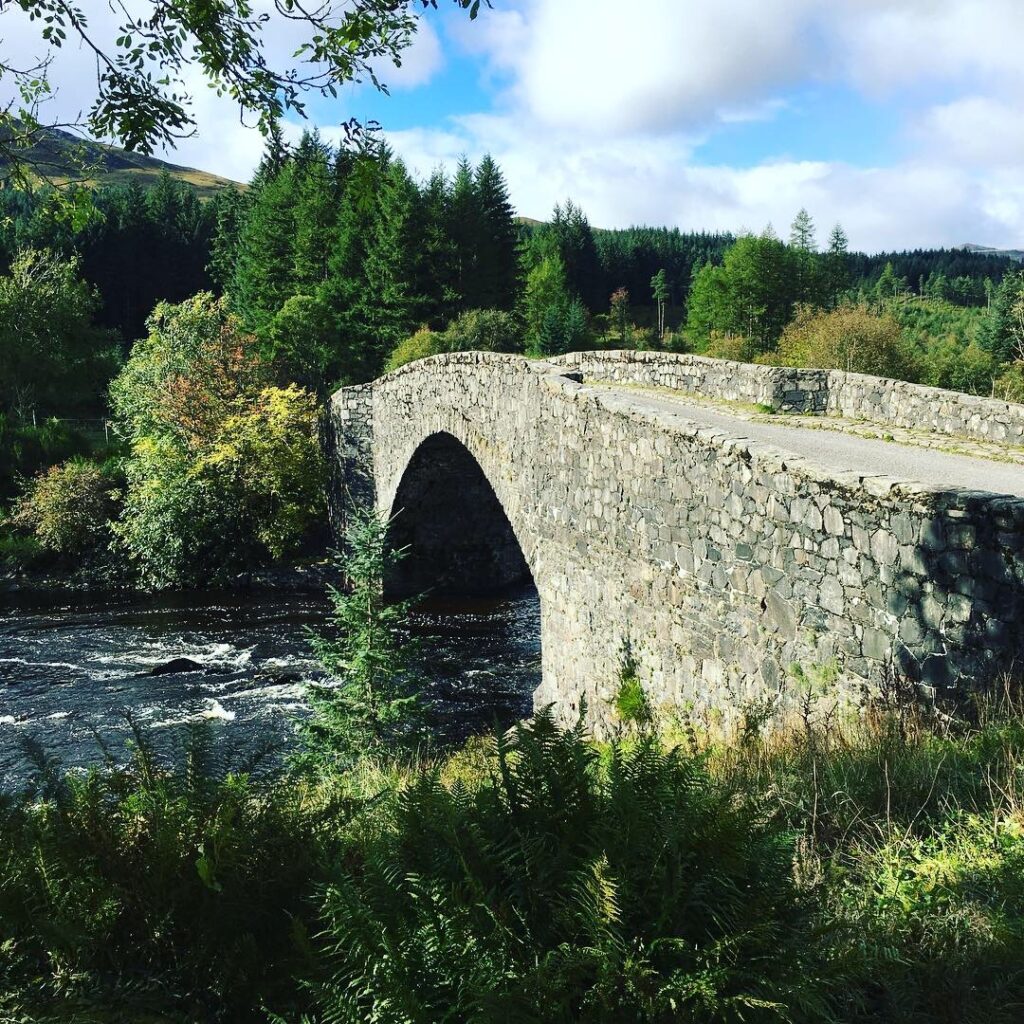 Traditional stone bridge with a large central arch crossing the flowing River Orchy in the Scottish Highlands, surrounded by dense evergreen trees, ferns and green hills beneath a partly cloudy blue sky.