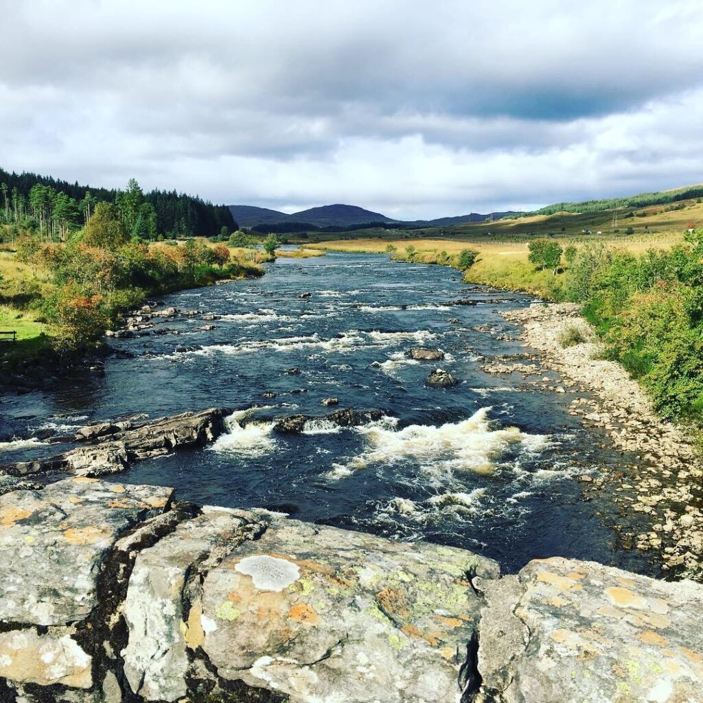 View of the River Orchy from a stone bridge near the start of the Bridge of Orchy to Fort William section of the West Highland Way, with water flowing over rocky rapids, surrounded by green woodland, open countryside and distant Scottish hills.