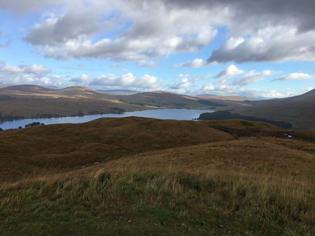 Panoramic view of a Highland loch along the Bridge of Orchy to Fort William section of the West Highland Way, surrounded by rolling brown and green hills, open moorland and distant mountains beneath a dramatic sky filled with clouds.
