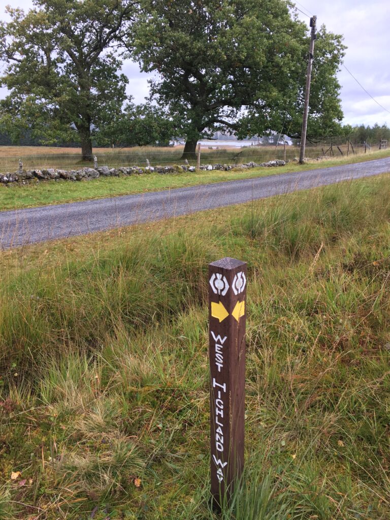 Close-up of a brown wooden West Highland Way waymarker featuring white trail symbols and yellow directional arrows, surrounded by long grass beside a narrow tarmac road, with mature trees, a low stone wall and a cloudy sky in the background.