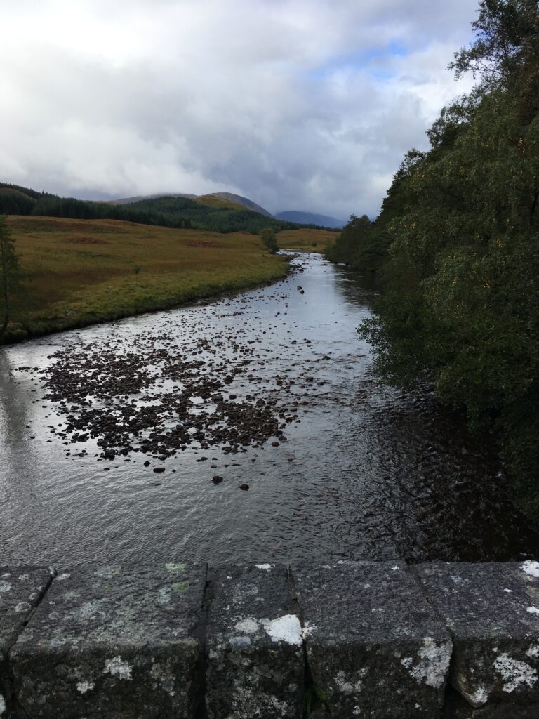 View of a wide, shallow river along the Bridge of Orchy to Fort William section of the West Highland Way, with scattered rocks beneath the flowing water, grassy moorland on one bank, dense woodland on the other and distant Highland hills beneath a cloudy sky.