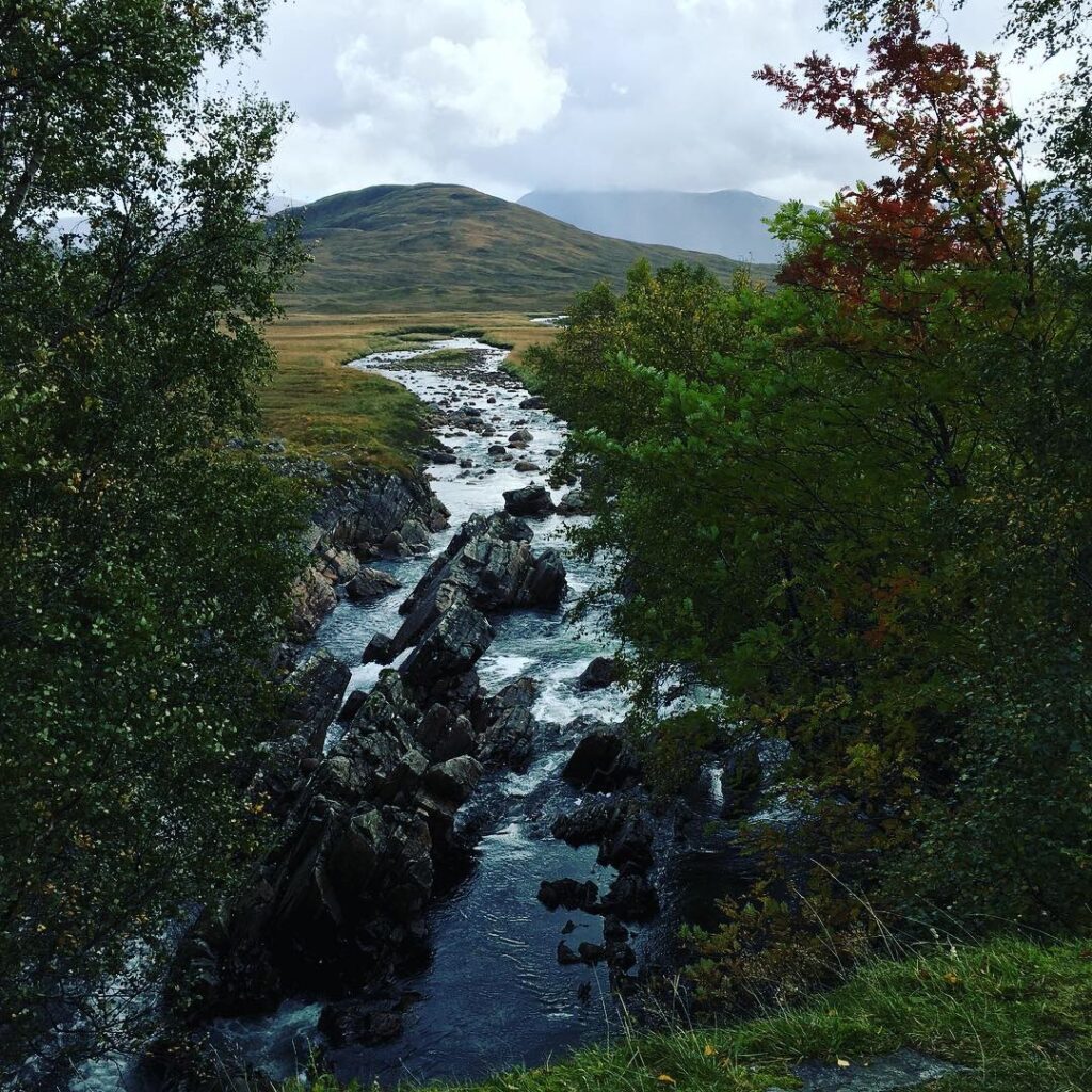 Scenic Scottish Highland river cascading between large jagged rocks and tree-lined banks before winding across open grassy moorland towards rolling green mountains, with distant peaks partially obscured by low clouds.