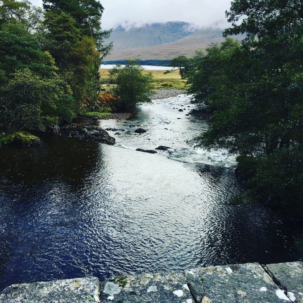 Scenic river flowing through a wooded valley along the Bridge of Orchy to Fort William section of the West Highland Way, with rocky banks, lush green trees, a distant loch and towering Scottish mountains partially obscured by low clouds.
