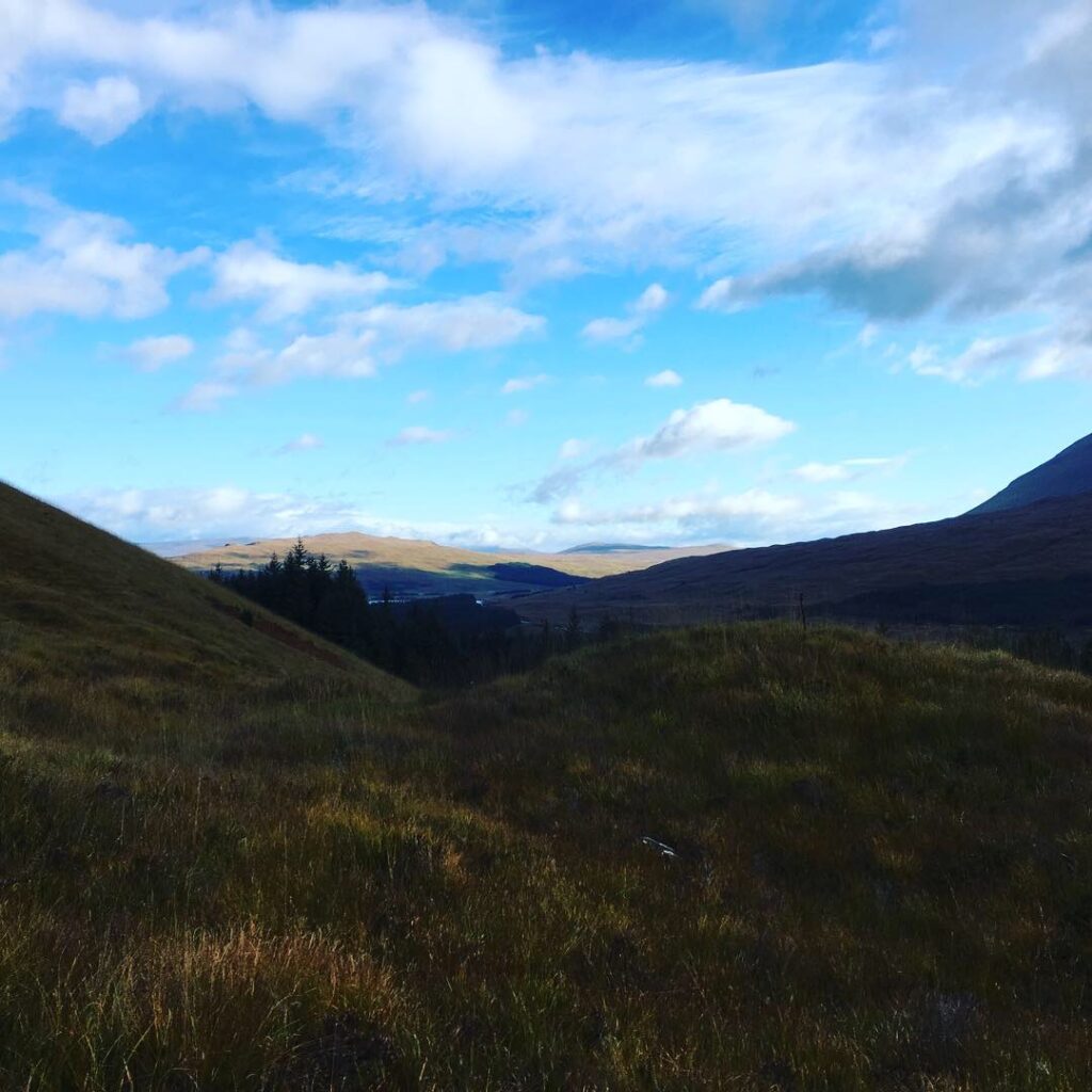 Expansive Highland valley along the Bridge of Orchy to Fort William section of the West Highland Way, with grassy hillsides, dark evergreen trees and distant sunlit mountains beneath a bright blue sky filled with scattered white clouds.