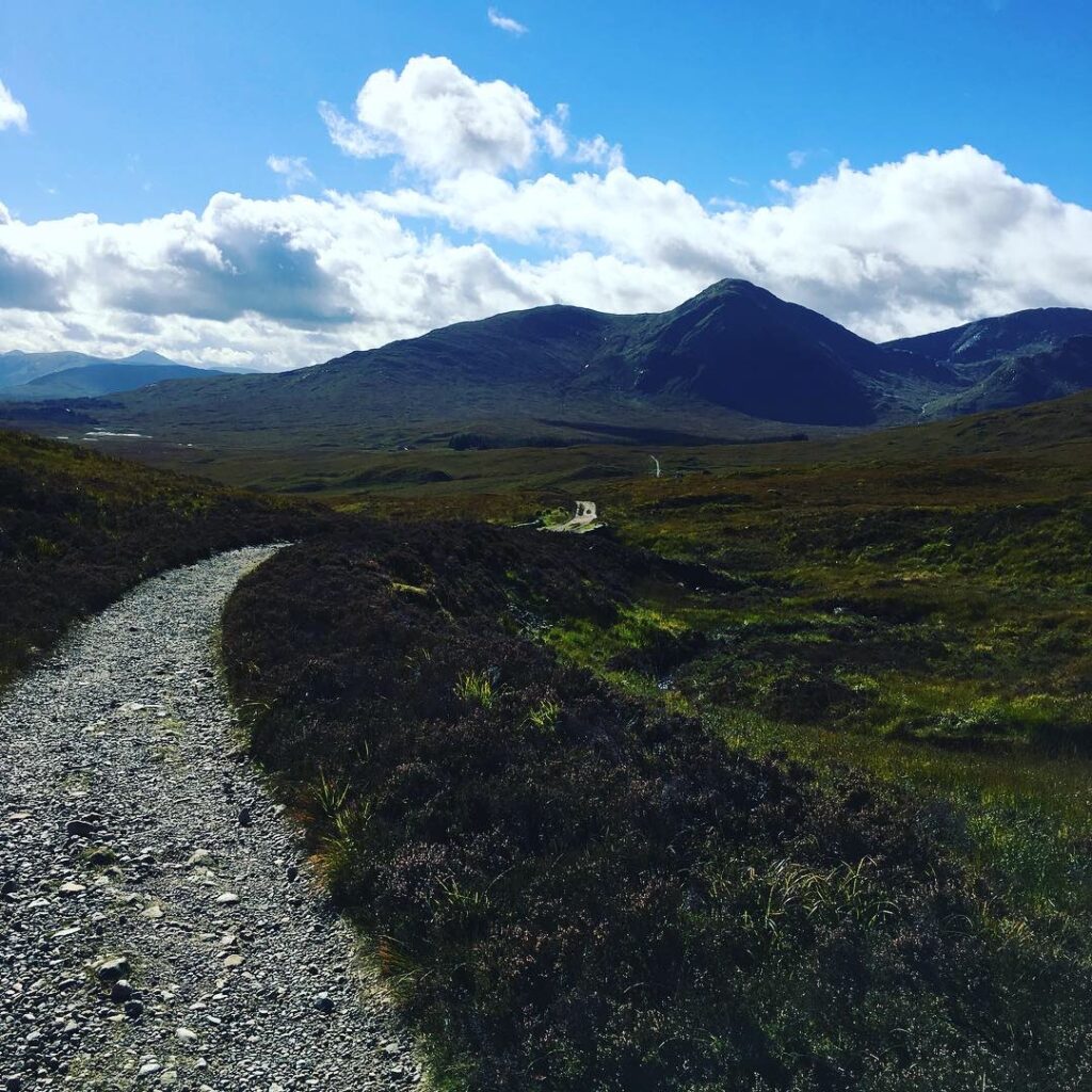 Rocky walking trail on the Bridge of Orchy to Fort William section of the West Highland Way, winding through open Highland moorland towards imposing mountain peaks beneath a blue sky dotted with white clouds.