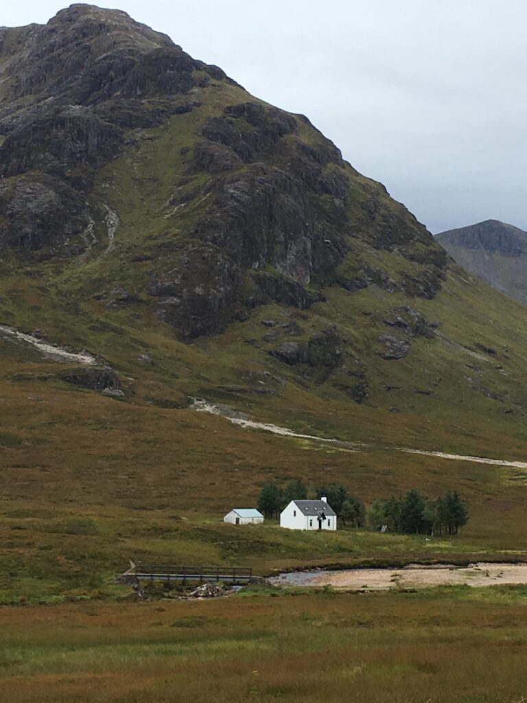 Isolated white cottage surrounded by open brown and green moorland beneath the towering, rocky slopes of a mountain in Glencoe, Scotland, with a small wooden footbridge, winding stream and scattered trees adding to the dramatic Highland landscape.