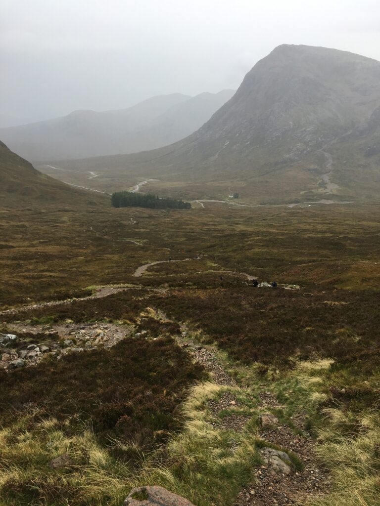 Winding rocky hiking trail on the Bridge of Orchy to Fort William section of the West Highland Way, overlooking a vast Highland valley surrounded by towering mountains partially obscured by mist and low cloud, with walkers visible along the path.