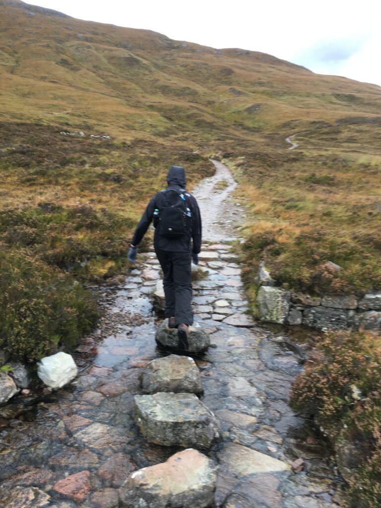 Hiker wearing black waterproof clothing and a backpack crossing a shallow stream on stepping stones along the Bridge of Orchy to Fort William section of the West Highland Way, with a wet, rocky mountain trail winding uphill through brown and green Highland moorland.