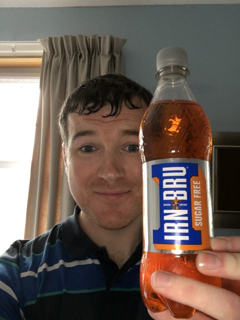 Close-up selfie of a smiling hiker holding a large bottle of Irn-Bru, Scotland’s famous orange soft drink, after a day of hiking in the Scottish Highlands, with a curtained window and blue walls in the background.
