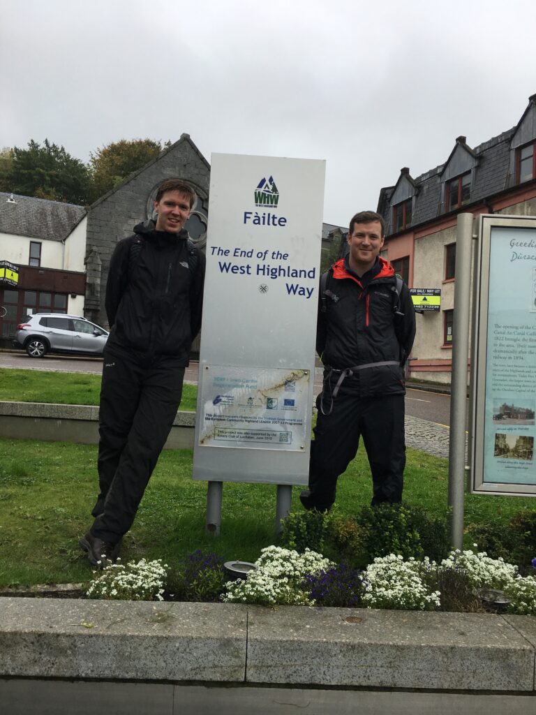 Two hikers celebrating the completion of the Bridge of Orchy to Fort William section of the West Highland Way, standing on either side of the official finishing sign in Fort William, Scotland, wearing waterproof hiking jackets and backpacks after their three-day walking adventure.