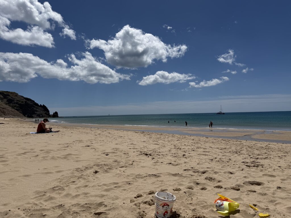 Golden sands stretching towards the Atlantic Ocean at Praia da Luz beach in Portugal, with colourful buckets and spades in the foreground, people paddling in the sea and a sailing boat offshore beneath a blue sky dotted with white clouds.