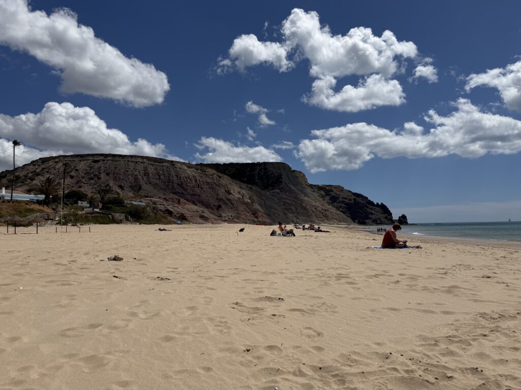 Wide sandy beach at Praia da Luz, with towering rocky cliffs, scattered sunbathers and the Atlantic Ocean beneath a blue sky during our week in the Algarve with a 3 year old.