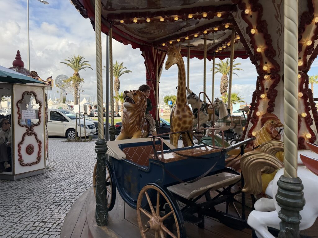 Decorative children's carousel beside Lagos Marina in Portugal, featuring a vintage blue carriage surrounded by animal figures including a giraffe, lions and horses, with glowing lights, palm trees and parked cars in the background.