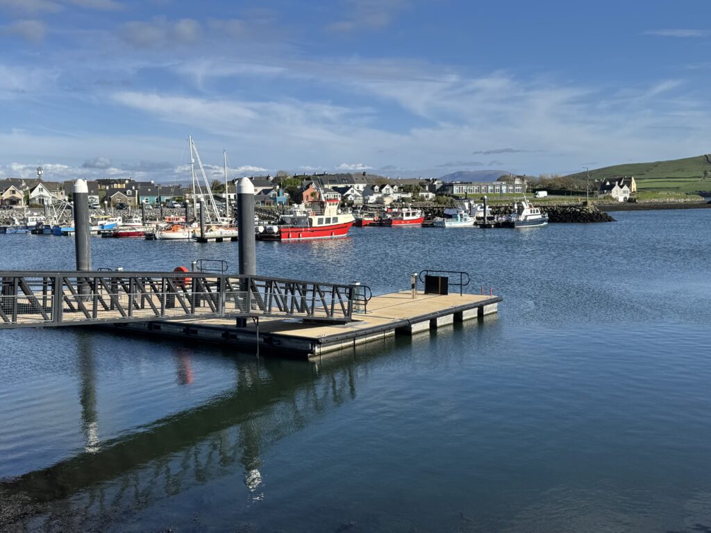 Scenic view of Dingle Harbour, featured in our guide to the best walks in Dingle, showing colourful fishing boats, a floating marina pontoon and waterfront buildings, with green hills in the background.