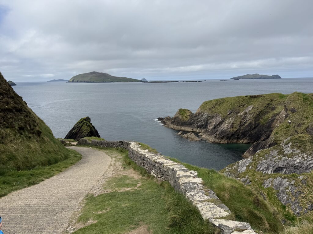 A narrow, winding pathway bordered by an old stone wall descends towards Dunquin Pier, overlooking a rocky coastal inlet surrounded by steep grassy cliffs, with the Blasket Islands visible across the Atlantic Ocean beneath cloudy skies.