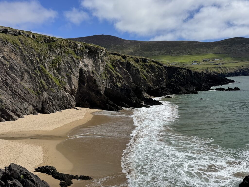 Elevated view of Coumeenoole Beach on the Dingle Peninsula, showing a small sandy cove sheltered by steep, rugged cliffs, with turquoise Atlantic waters washing onto the shore and green hills beneath a partly cloudy blue sky.
