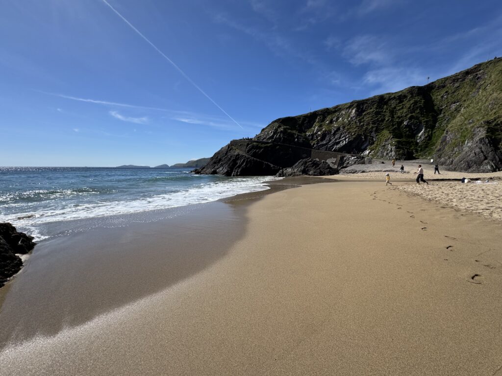 Ground-level view of Coumeenoole Beach on the Dingle Peninsula, showing a sweeping sandy cove, waves washing onto the shore, visitors exploring the beach and steep, grassy cliffs rising above the rocky coastline beneath a clear blue sky.