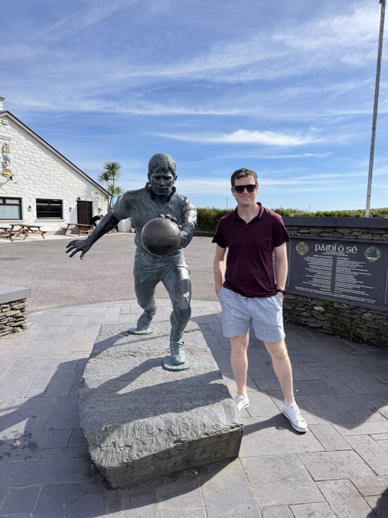 A man wearing sunglasses, a burgundy polo shirt and light blue shorts standing beside the bronze statue of Kerry football legend Páidí Ó Sé holding a football, outside the family pub in Ventry, County Kerry, on a sunny day.