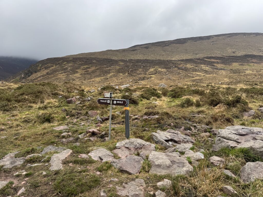 Kerry Camino signpost pointing towards Tralee and Dingle, surrounded by rocky moorland with the lower slopes of the Slieve Mish Mountains behind.