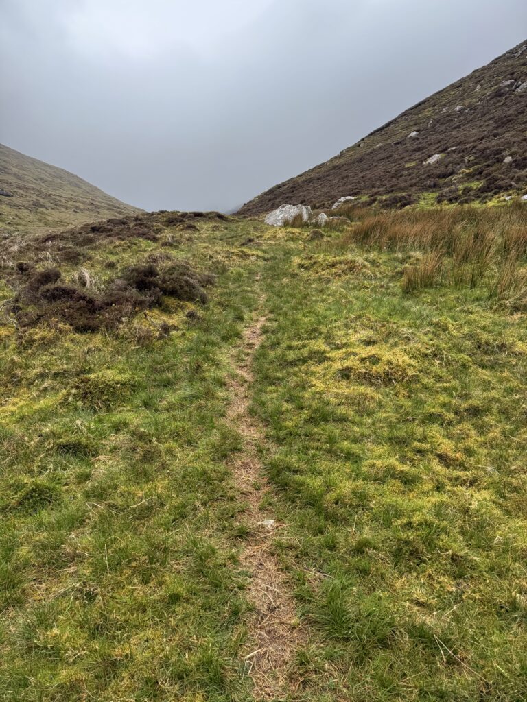 Narrow grassy mountain path leading through Derrymore Glen, with steep slopes on both sides and low cloud obscuring the higher ground ahead.