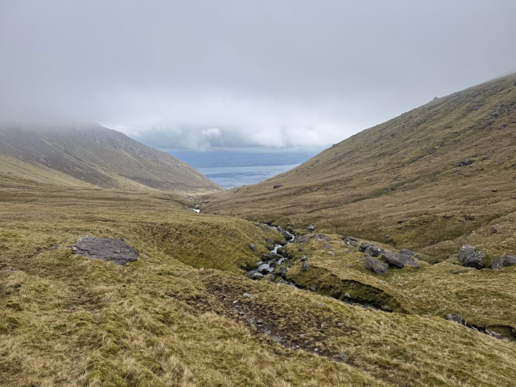View back through Derrymore Glen, with a rocky river winding between steep mountain slopes towards the distant Kerry coastline beneath low cloud.