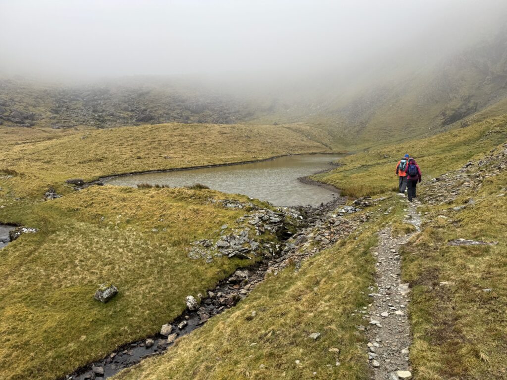 Two hikers following the Caherconree and Baurtregaum Loop past a small mountain lake in Derrymore Glen, with a rocky path, stream and low cloud covering the surrounding slopes.