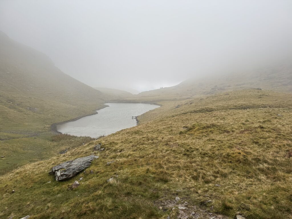 Derrymore Lough surrounded by steep grassy mountain slopes, with thick mist and low cloud hiding the higher ground above the lake.