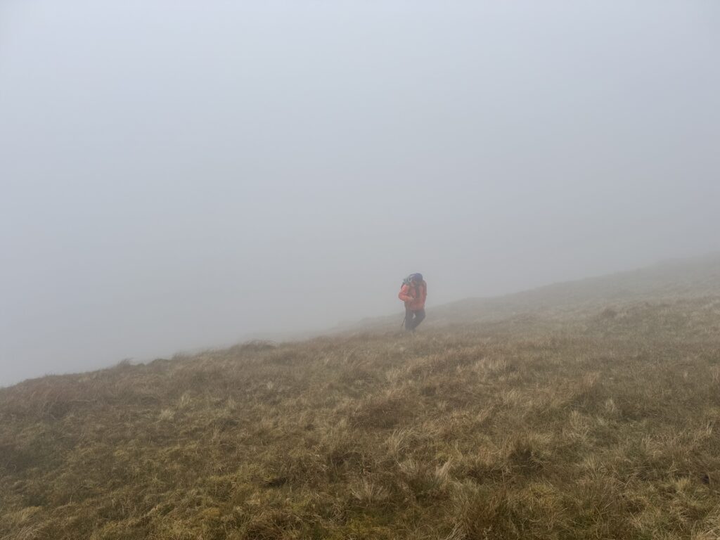 Lone hiker climbing a steep grassy mountain slope in dense fog, with visibility reduced to only a short distance ahead.