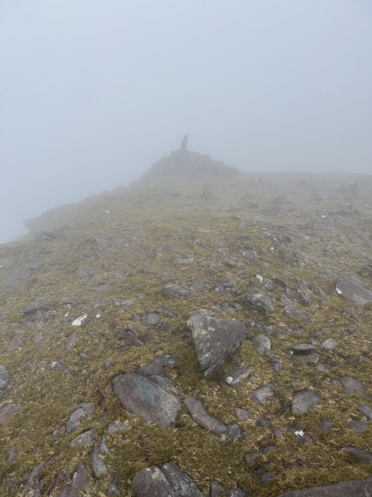 Caherconree summit on the Caherconree and Baurtregaum Loop, with a rocky ridge and summit marker emerging through thick fog and very poor visibility.
