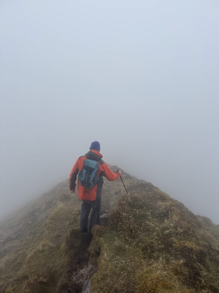 Hiker walking along a narrow ridge on the Caherconree and Baurtregaum Loop, surrounded by dense mist with steep drops disappearing into the cloud on either side.