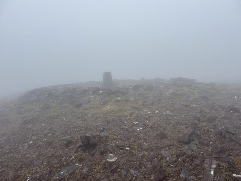 Baurtregaum summit in thick fog, with a concrete trig pillar standing among rocks and very limited visibility across the mountain top.