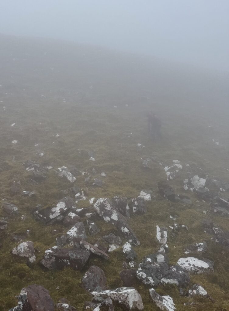 Hiker descending a steep, rock-strewn mountain slope in thick mist, with uneven ground and visibility reduced to only a short distance ahead.
